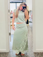 A woman takes a mirror selfie in a boutique wearing a sage green two-piece set consisting of a sleeveless crop top and a matching long skirt with lace trim.