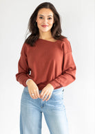 A woman modeling a rust-colored, boat-neck long-sleeve sweatshirt paired with light blue jeans against a plain white studio background.