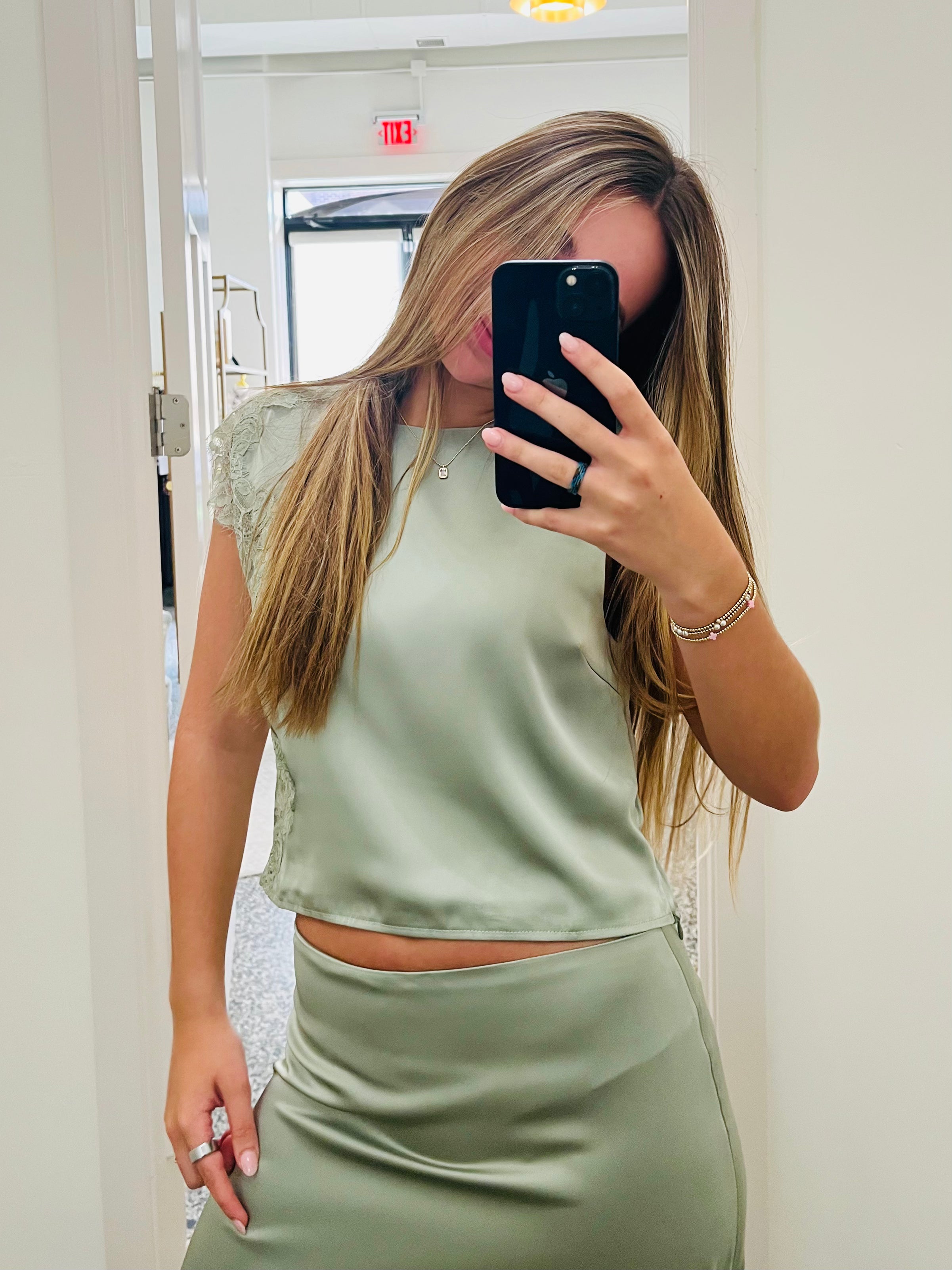 A woman takes a mirror selfie in a fitting room wearing a sage green satin crop top and matching high-waisted satin skirt.