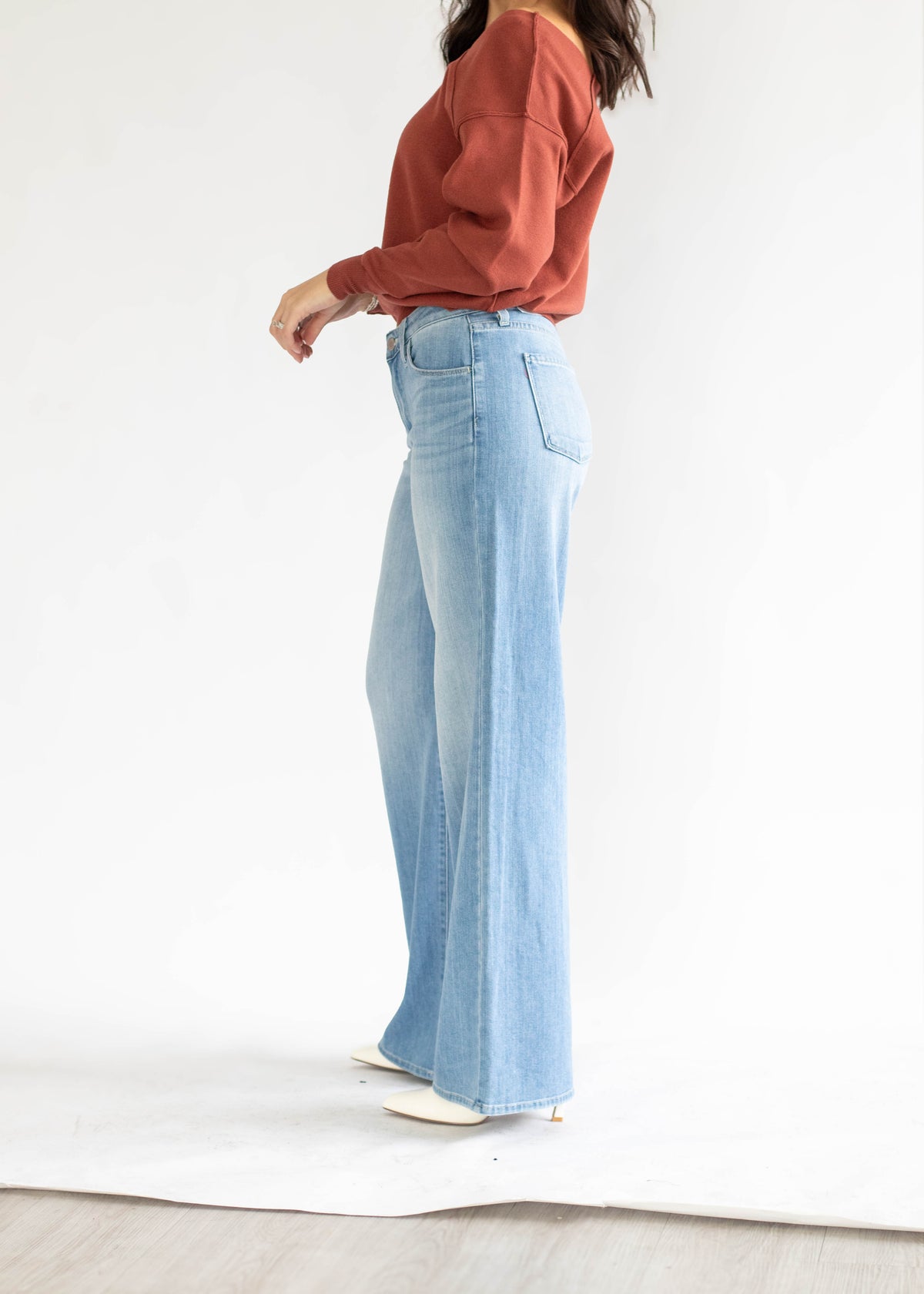 A model wearing a rust-colored long-sleeve top and light-wash wide-leg denim jeans, standing in profile against a white studio background.
