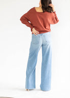A woman shown from the back wearing a rust-colored, wide-neck long-sleeve sweatshirt and light-wash wide-leg denim jeans against a plain white studio background.