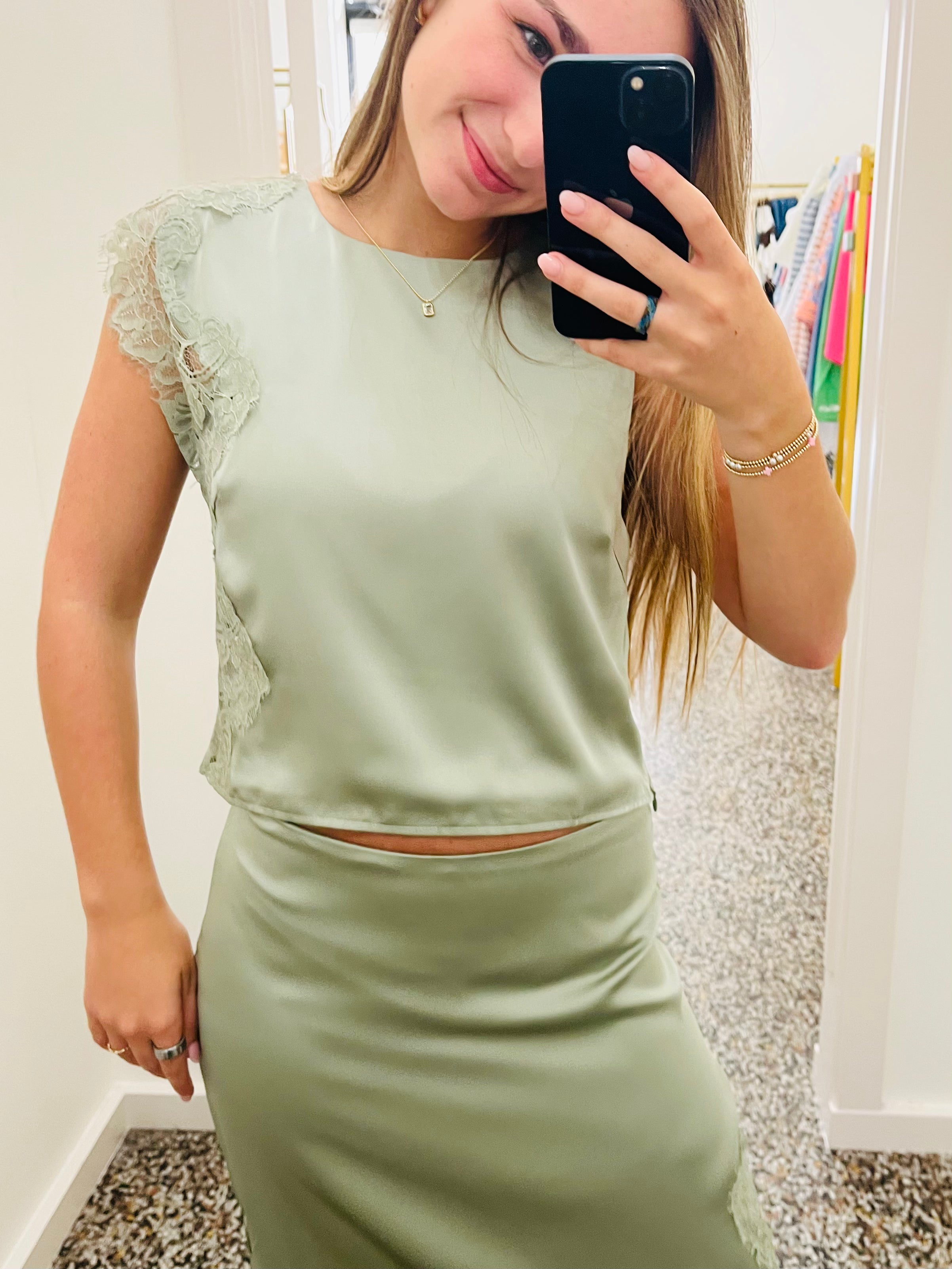 A woman takes a mirror selfie wearing a sage green satin two-piece set consisting of a short-sleeve crop top with lace detailing on the sleeves and a matching high-waisted pencil skirt.
