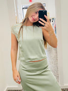 A woman takes a mirror selfie in a boutique wearing a sage green satin two-piece set consisting of a sleeveless crop top with lace detailing on the shoulders and a matching high-waisted pencil skirt.
