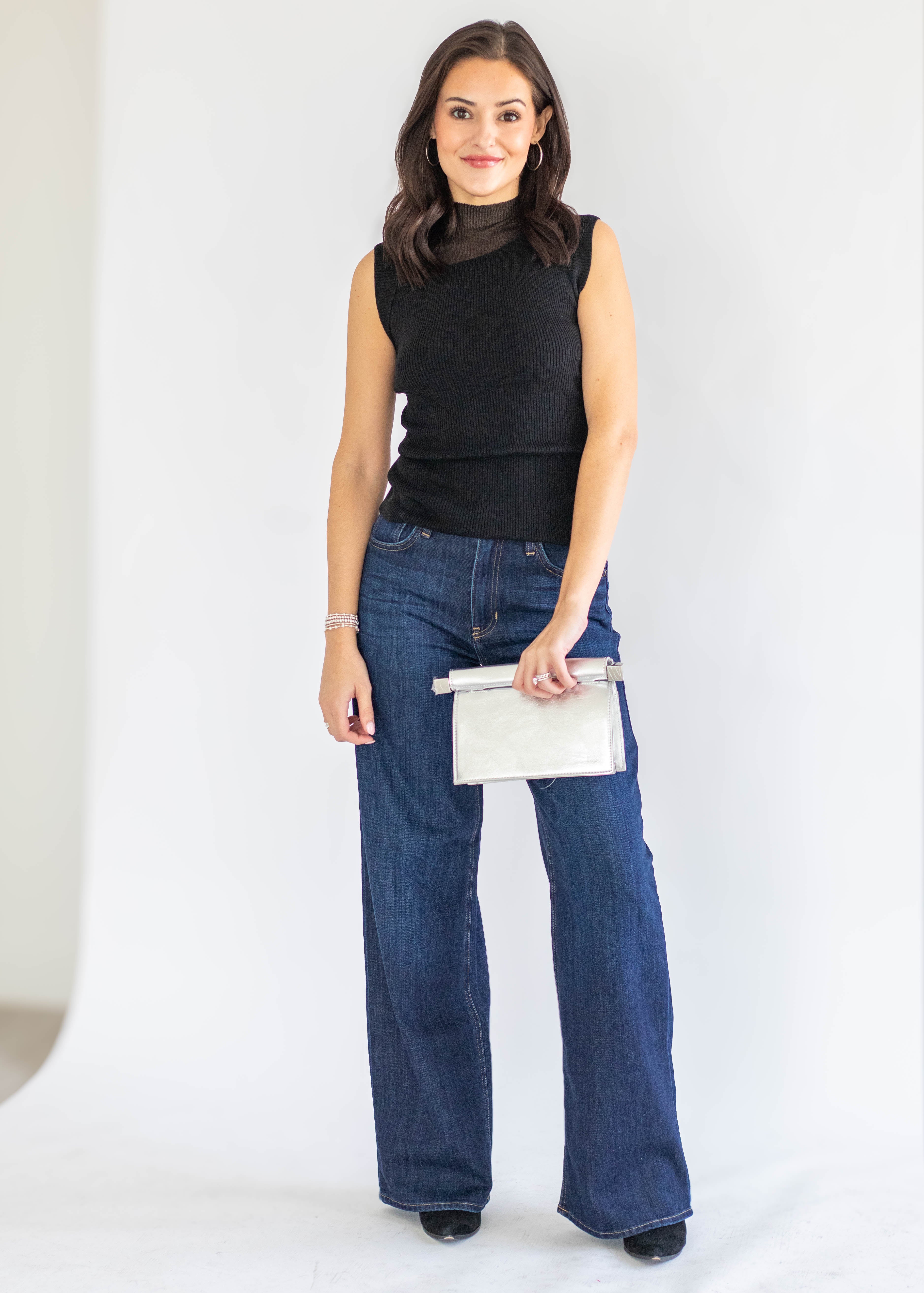 A woman modeling a sleeveless black mock neck top and dark blue wide-leg jeans, holding a small white clutch bag against a plain white studio background.