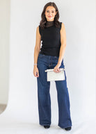 A woman modeling a sleeveless black mock neck top and dark blue wide-leg jeans, holding a small white clutch bag against a plain white studio background.