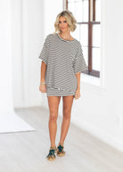 A woman modeling an oversized black and white horizontally striped t-shirt dress in a bright, modern room with white walls and a window.