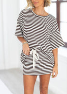 A woman wearing a brown and white horizontally striped short-sleeve tunic dress with a white drawstring tie at the waist, shown in a brightly lit indoor setting.