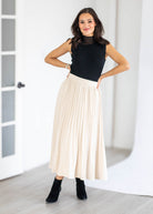 A woman modeling a black sleeveless mock-neck top and a high-waisted, pleated cream maxi skirt, paired with black ankle boots, in a bright, minimalist room.