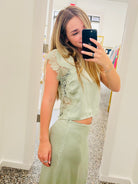A woman takes a mirror selfie in a boutique wearing a sage green two-piece set consisting of a short-sleeve satin crop top with lace detailing and a matching high-waisted satin skirt.