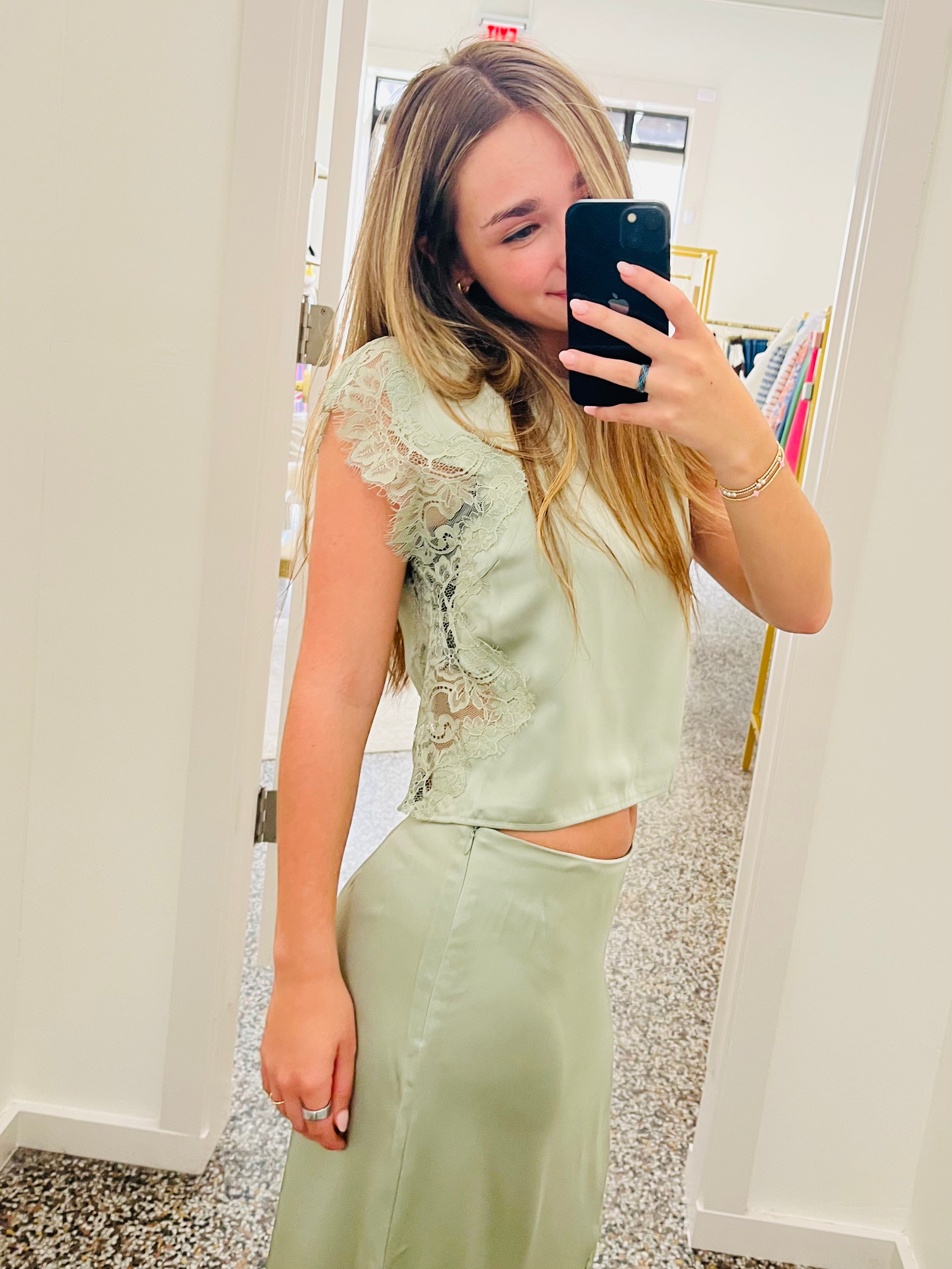 A woman takes a mirror selfie in a boutique wearing a sage green two-piece set consisting of a short-sleeve satin crop top with lace detailing and a matching high-waisted satin skirt.