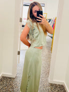 A woman takes a mirror selfie in a boutique wearing a sage green two-piece set consisting of a short-sleeve top with floral embroidery and a matching long skirt with a side cutout.