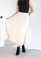 A woman wearing a black sleeveless top and a high-waisted, pleated cream-colored midi skirt, paired with black ankle boots, standing in a bright room.