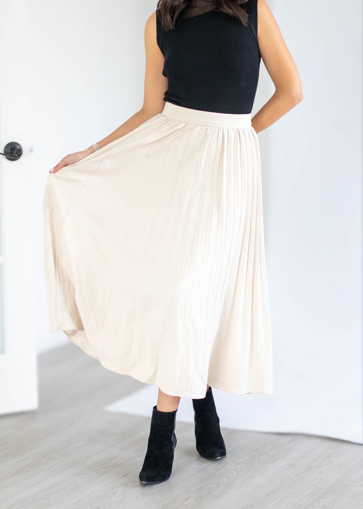 A woman wearing a black sleeveless top and a high-waisted, pleated cream-colored midi skirt, paired with black ankle boots, standing in a bright room.