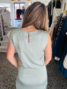 A woman viewed from behind wearing a sage green sleeveless dress with lace detailing on the shoulders, standing inside a clothing boutique.