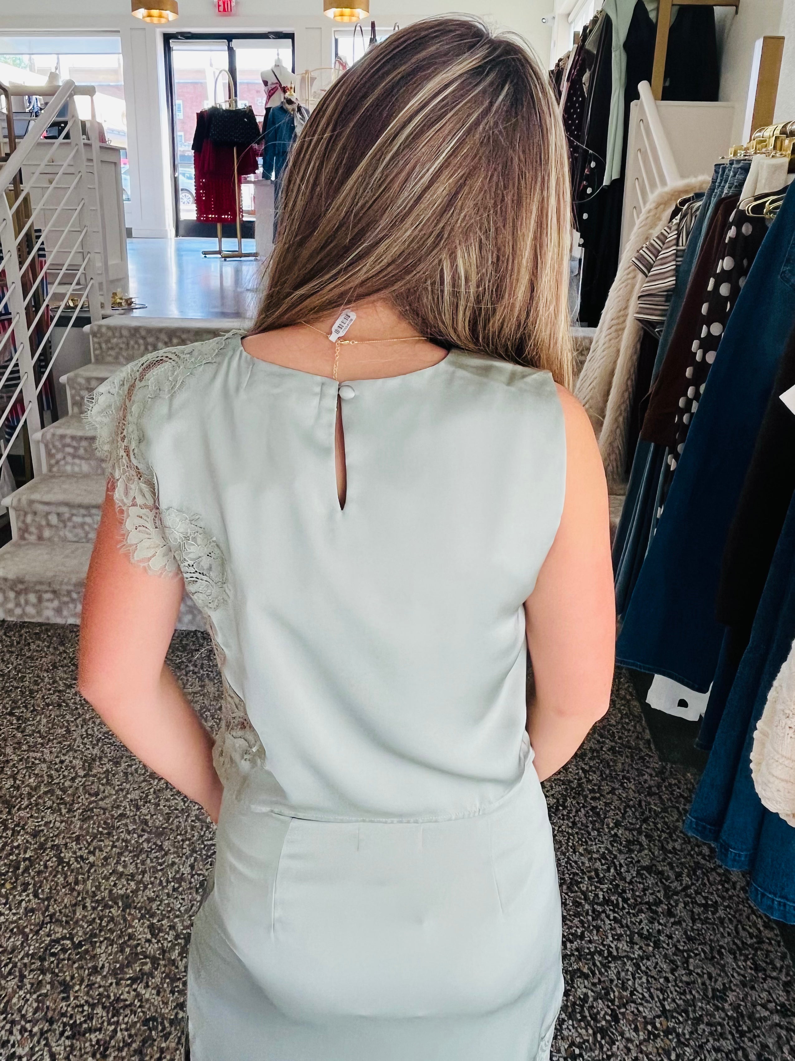 A woman viewed from behind wearing a sage green sleeveless dress with lace detailing on the shoulders, standing inside a clothing boutique.
