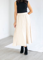 A woman wearing a black sleeveless top and a high-waisted, cream-colored pleated midi skirt, paired with black ankle boots, standing in a bright room.