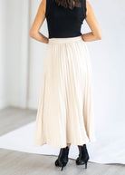 A woman shown from the back wearing a long pleated cream maxi skirt, a black sleeveless top, and black ankle boots in a bright indoor setting.