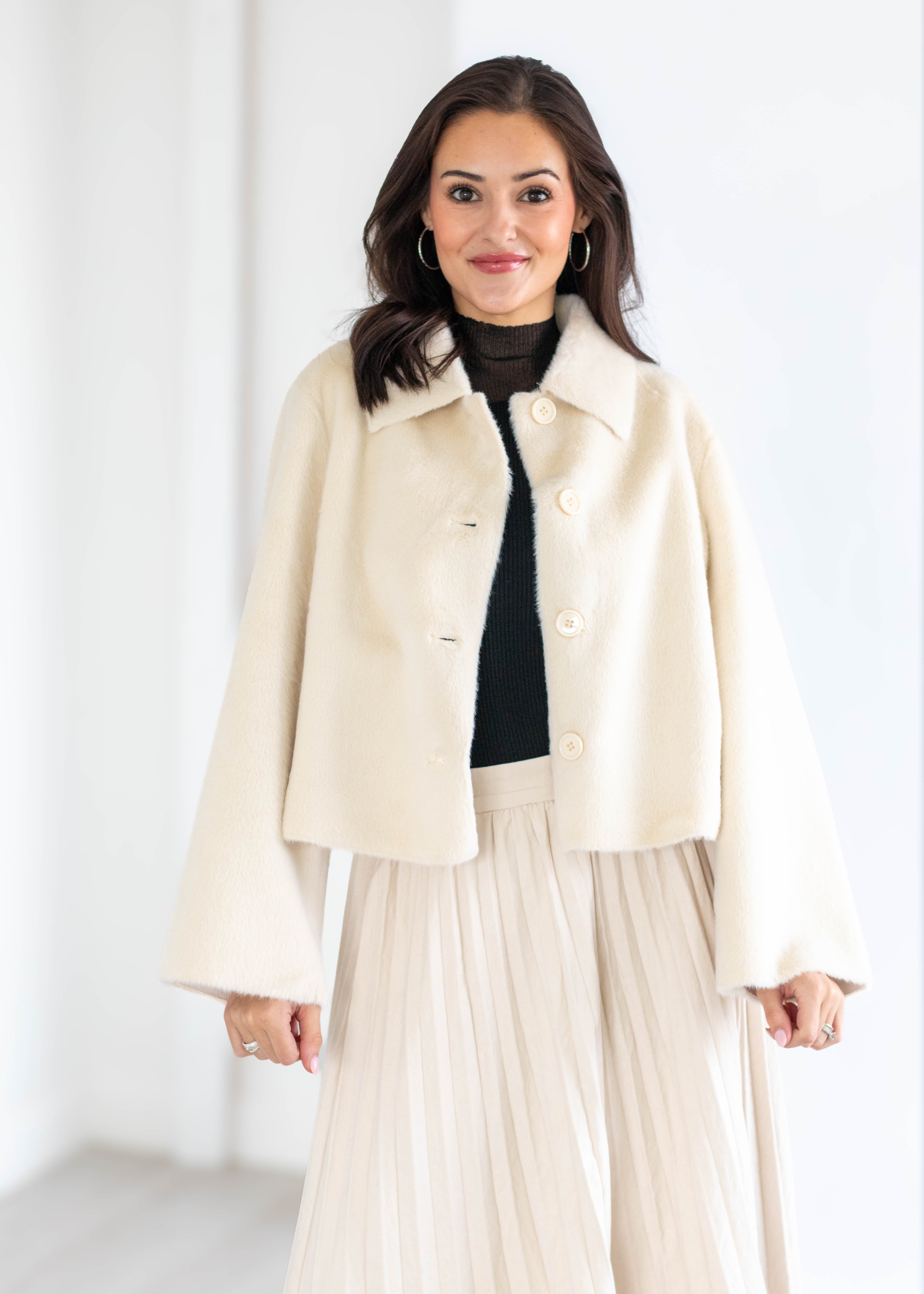 A woman modeling a cream-colored cropped jacket with a collar and large buttons, worn over a black turtleneck and a pleated cream skirt, set against a bright white indoor background.