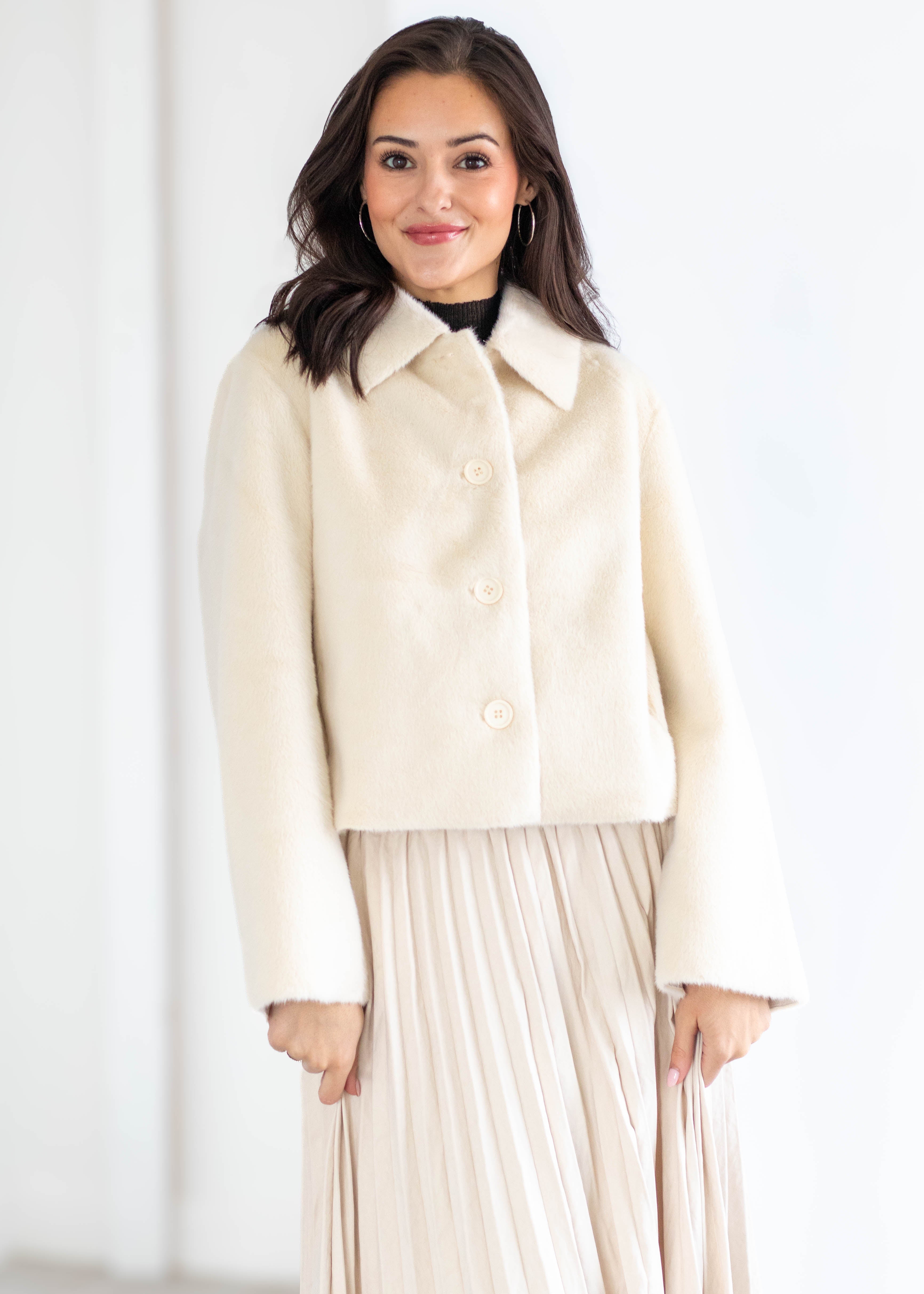 A woman modeling a cropped cream-colored faux fur jacket with a collar and three buttons, paired with a matching pleated skirt, against a bright white background.