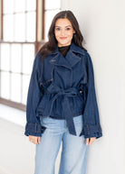 A woman modeling a dark blue denim belted jacket with light blue jeans, standing in front of a bright white wall with a window.
