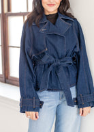 A woman wearing a dark blue denim belted jacket over a black top and light blue jeans, standing indoors near a window.