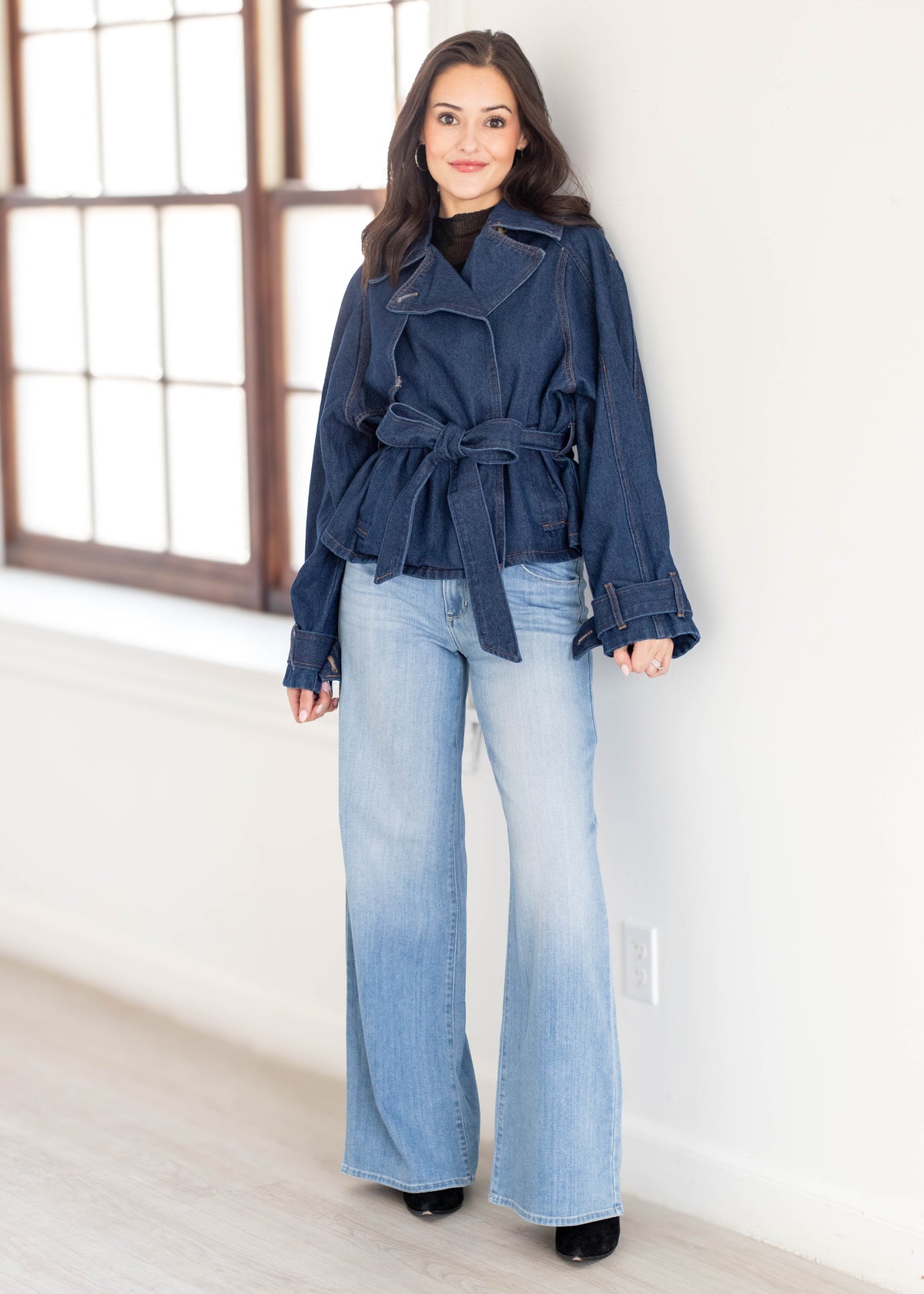 A woman modeling a dark blue belted denim jacket and light blue wide-leg denim jeans, standing in a bright room with a white wall and a large window.
