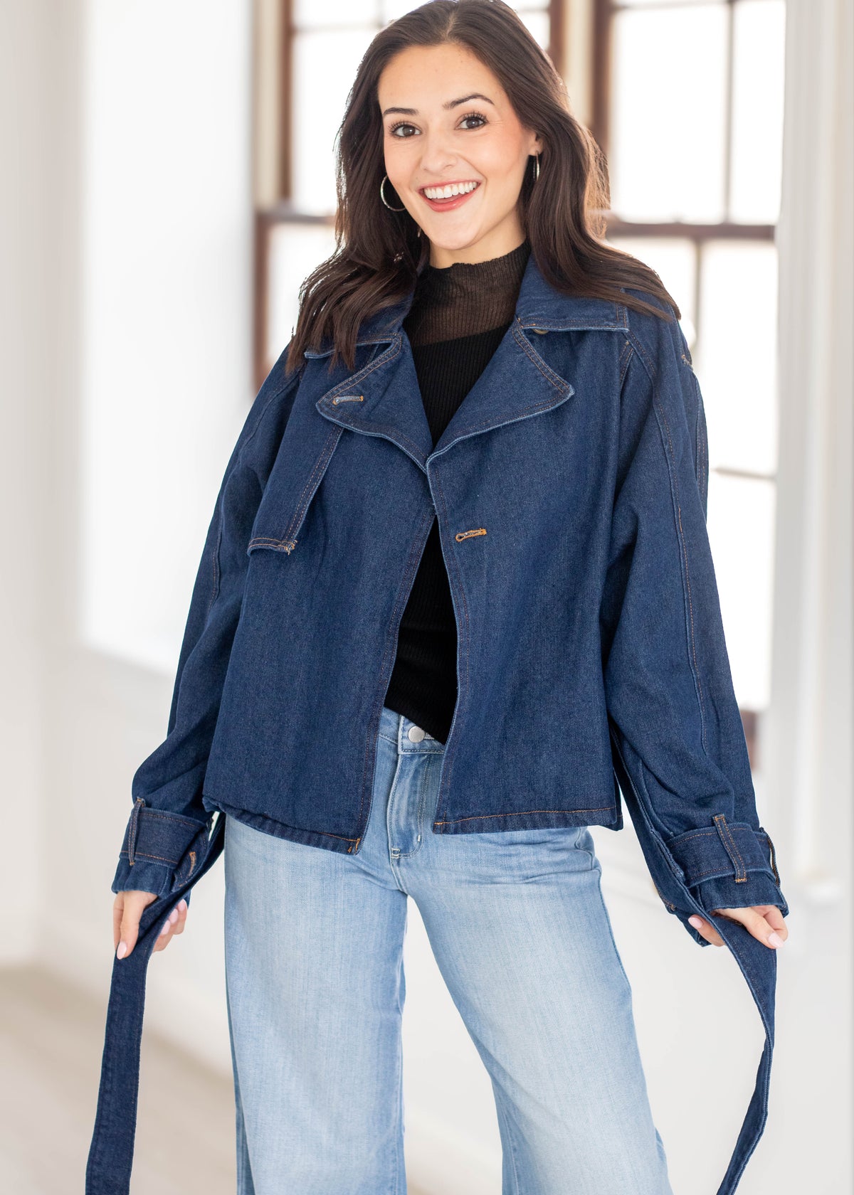 A woman wearing a dark blue denim jacket with a matching tie belt, styled over a black turtleneck and light blue jeans, standing in a bright indoor setting.