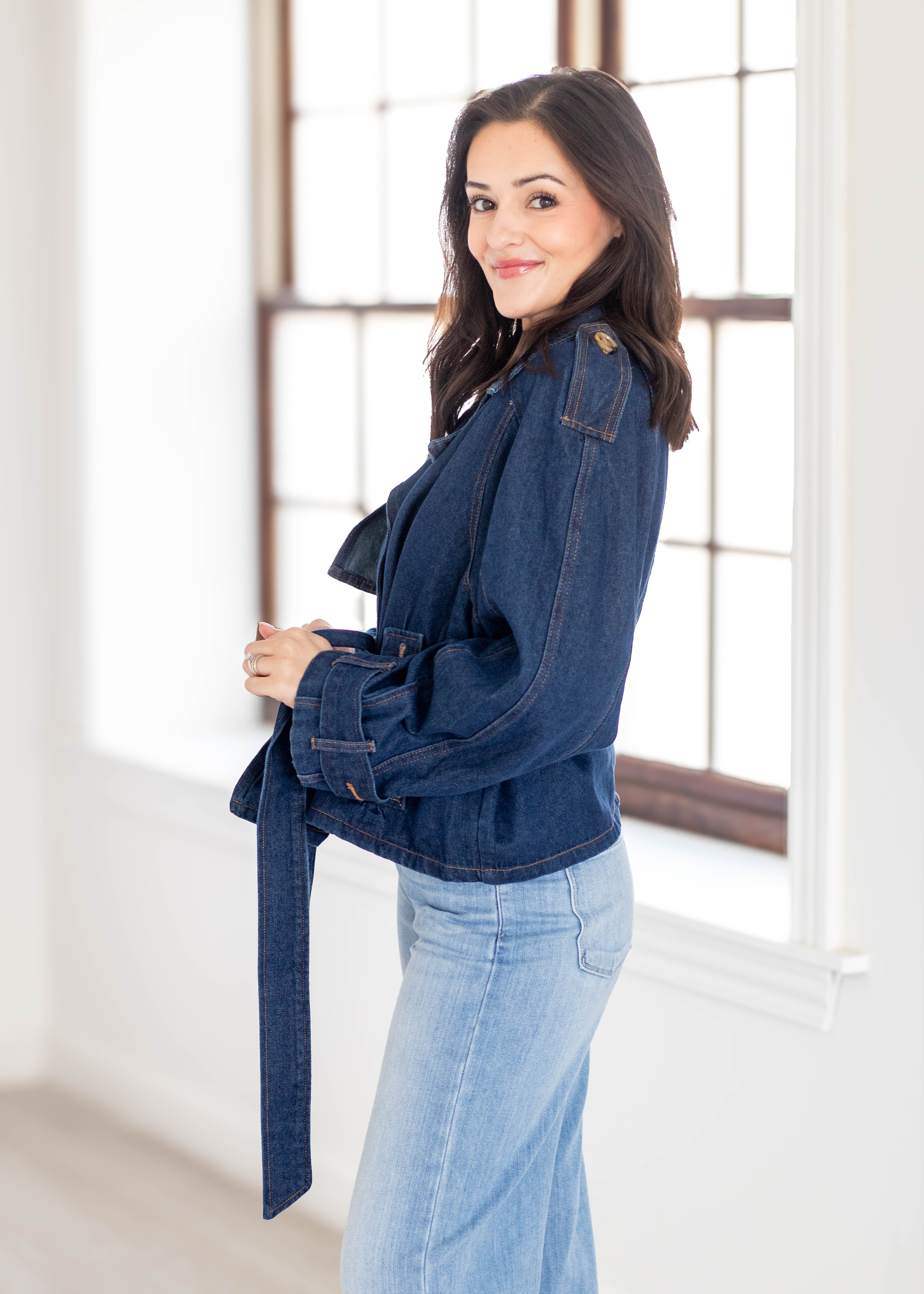 A woman wearing a dark blue denim belted jacket and light blue jeans, standing in a bright room with a large window.