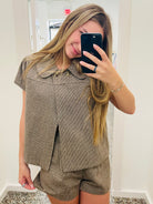 A woman takes a mirror selfie wearing a matching brown and beige houndstooth short-sleeve top and shorts set. She is holding a black smartphone to take the photo in what appears to be a boutique fitting room.