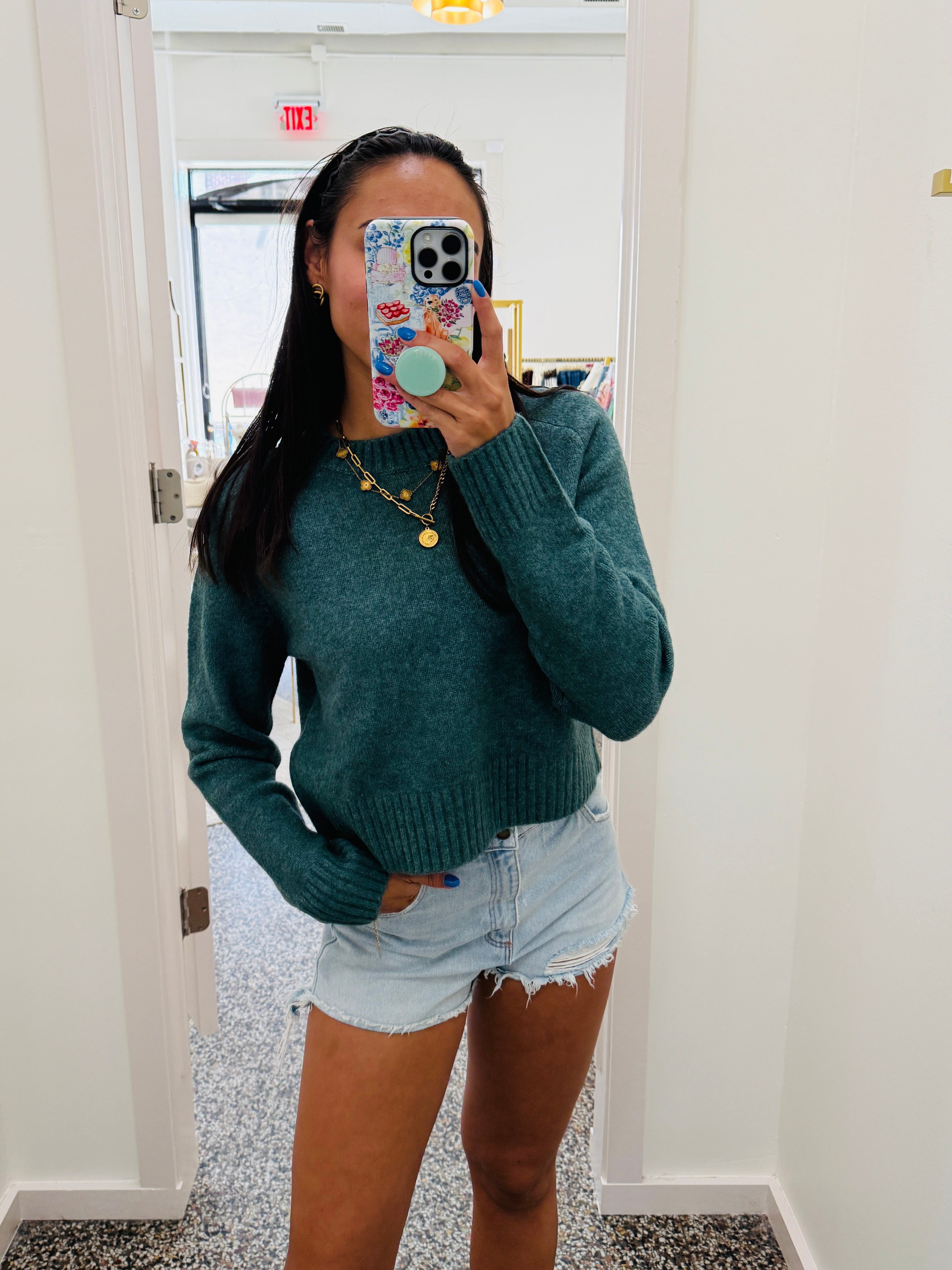 A woman takes a mirror selfie in a hallway, wearing a teal green knit sweater and light-wash denim shorts.
