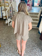 A woman viewed from behind wearing a matching taupe short-sleeve button-up shirt and shorts set in a clothing store.