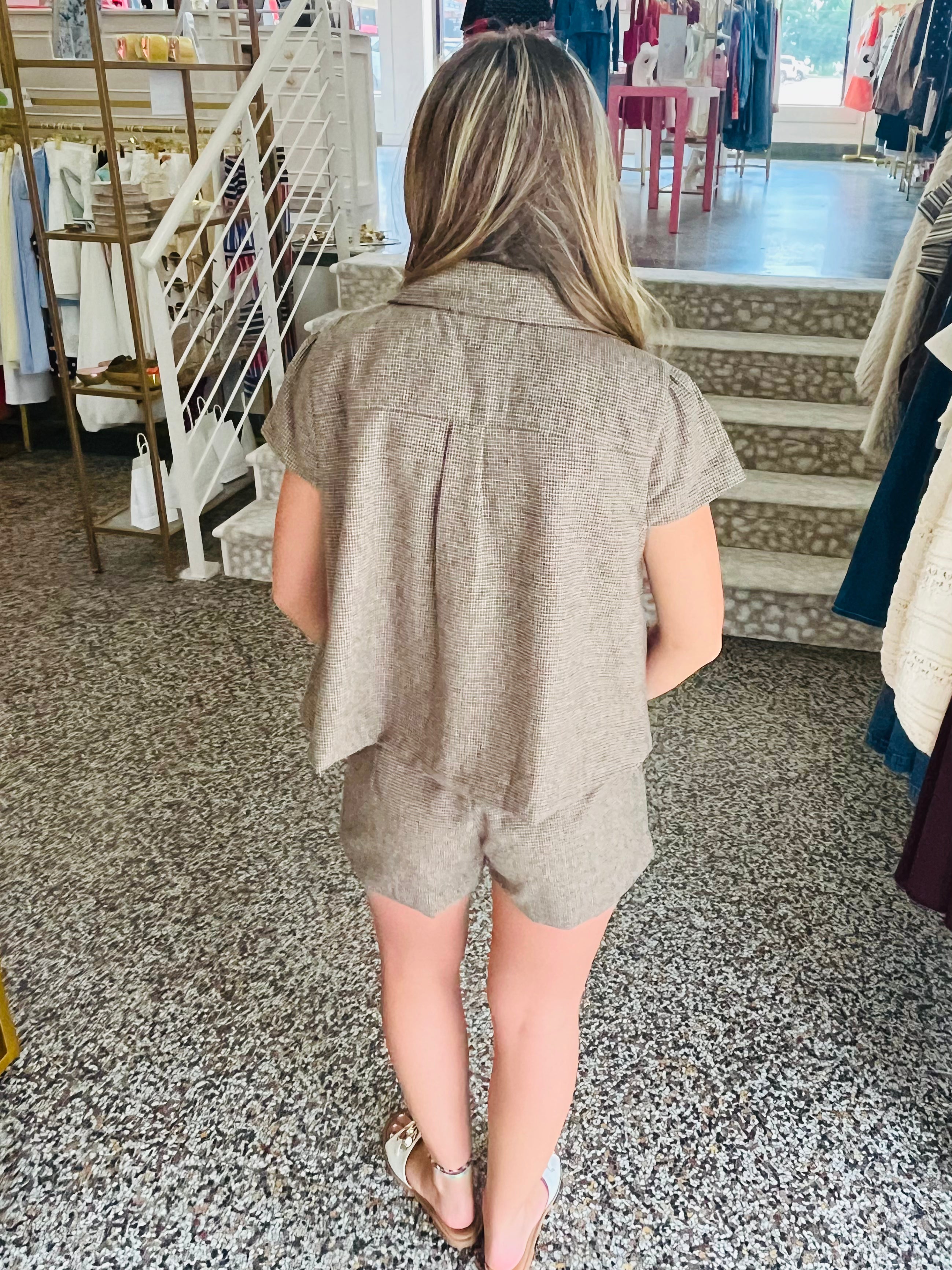 A woman viewed from behind wearing a matching taupe short-sleeve button-up shirt and shorts set in a clothing store.