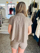 A woman viewed from behind wearing a textured taupe short-sleeve collared shirt and matching shorts in a clothing boutique.