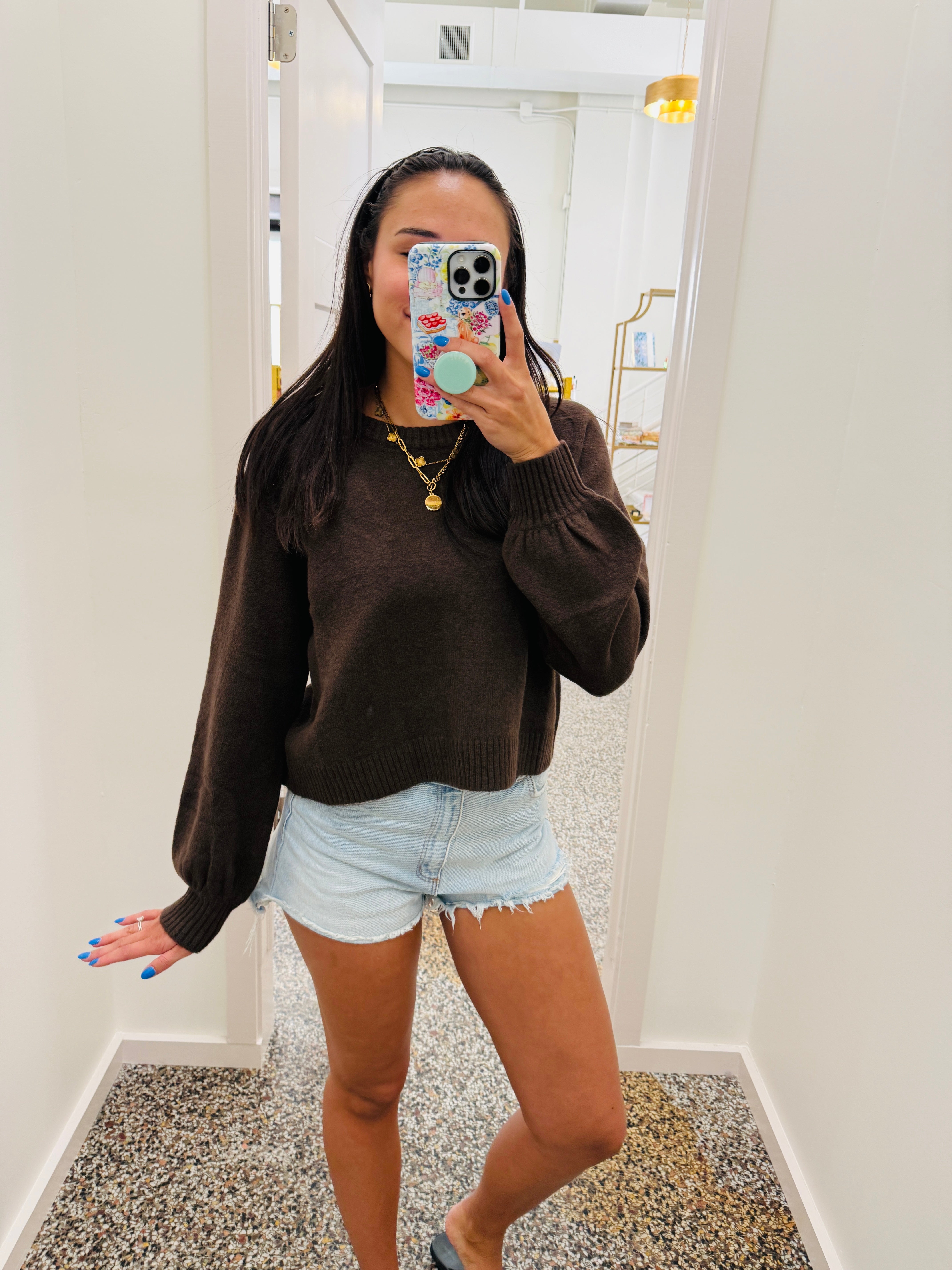 A woman takes a mirror selfie in a fitting room wearing a dark brown knit sweater and light blue denim shorts.