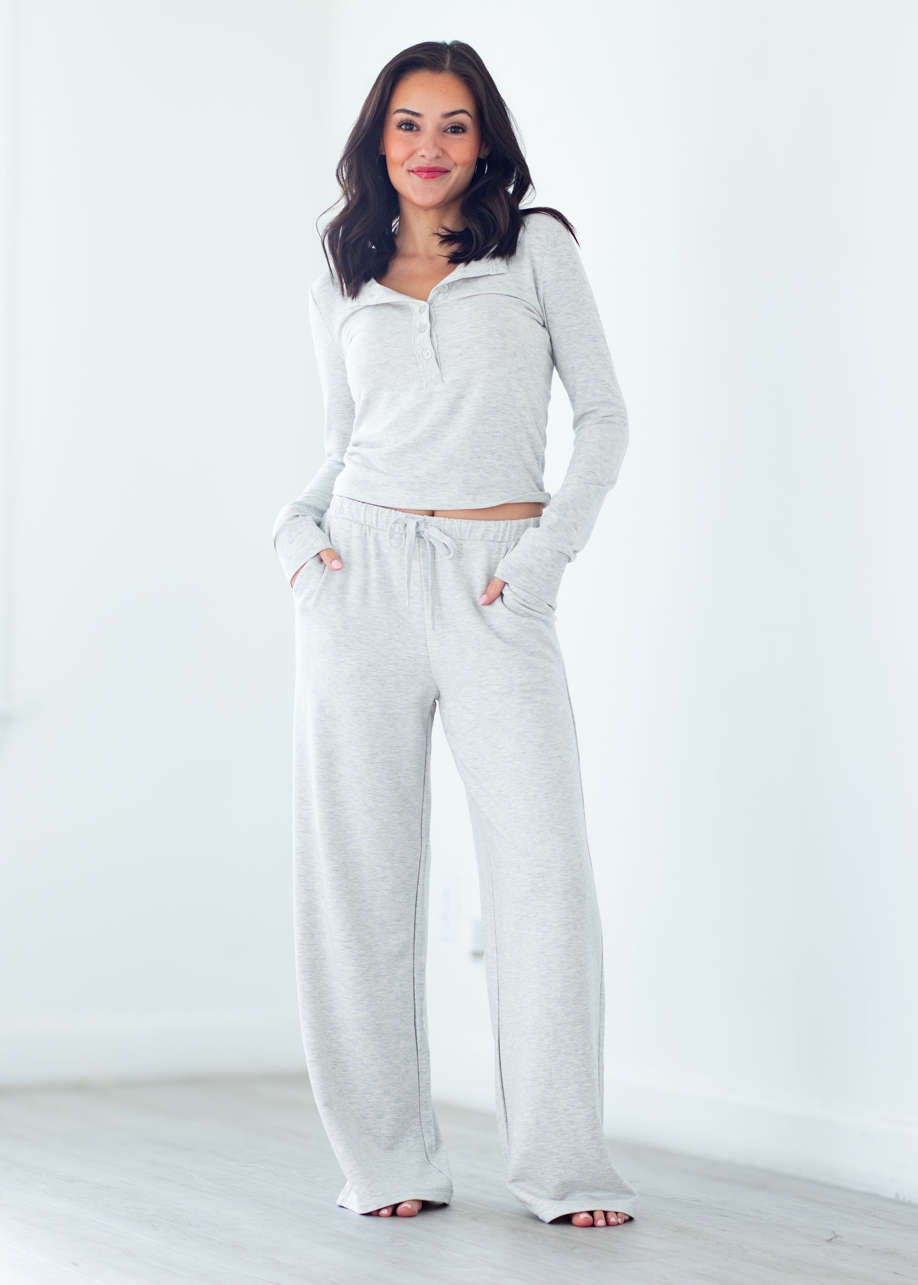 A woman modeling a light heather gray lounge set consisting of a long-sleeve half-zip top and matching wide-leg sweatpants, standing in a bright, minimalist room.