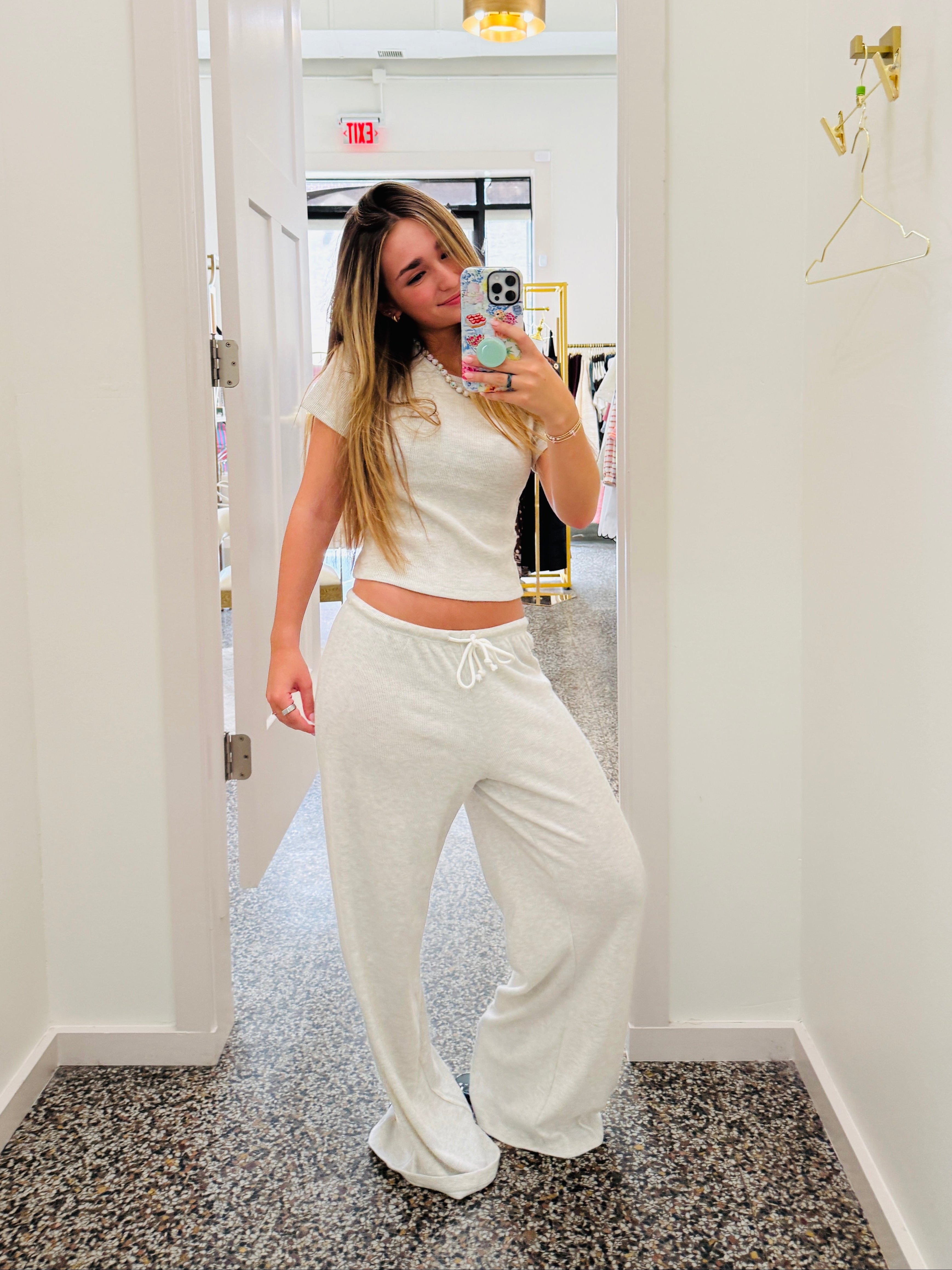 A woman takes a mirror selfie in a boutique fitting room wearing a matching cream two-piece lounge set consisting of a short-sleeve crop top and wide-leg sweatpants.
