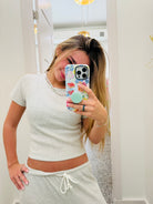 A woman taking a mirror selfie wearing a light gray short-sleeve crop top and matching gray sweatpants, holding a floral-patterned phone case with a mint green pop socket.
