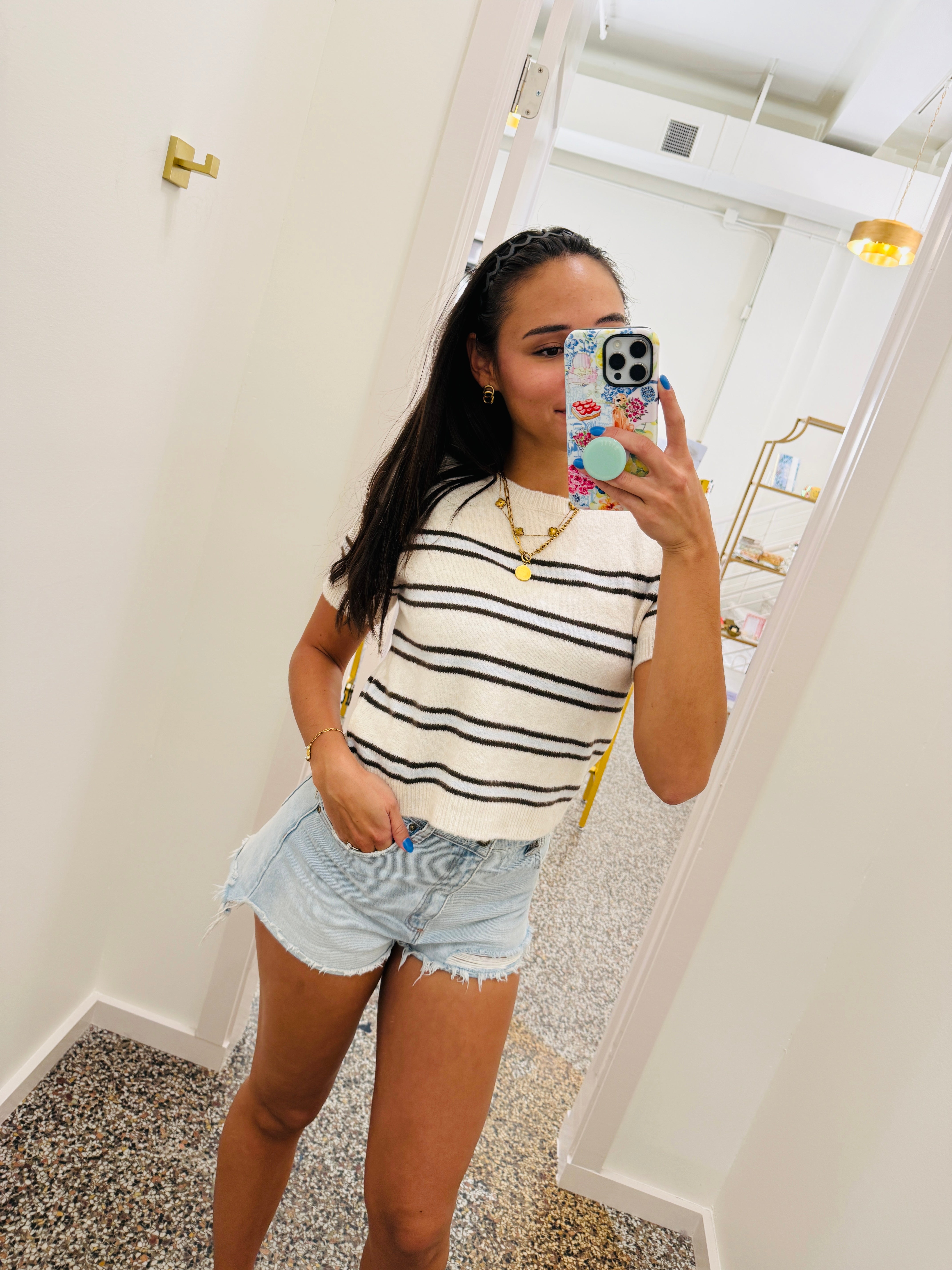 A woman takes a mirror selfie in a boutique wearing a white short-sleeve top with black horizontal stripes, light-wash denim shorts, and gold jewelry.