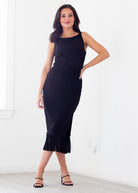 A woman modeling a sleeveless black midi dress with a draped neckline and a fringed hem, standing in a bright white room.