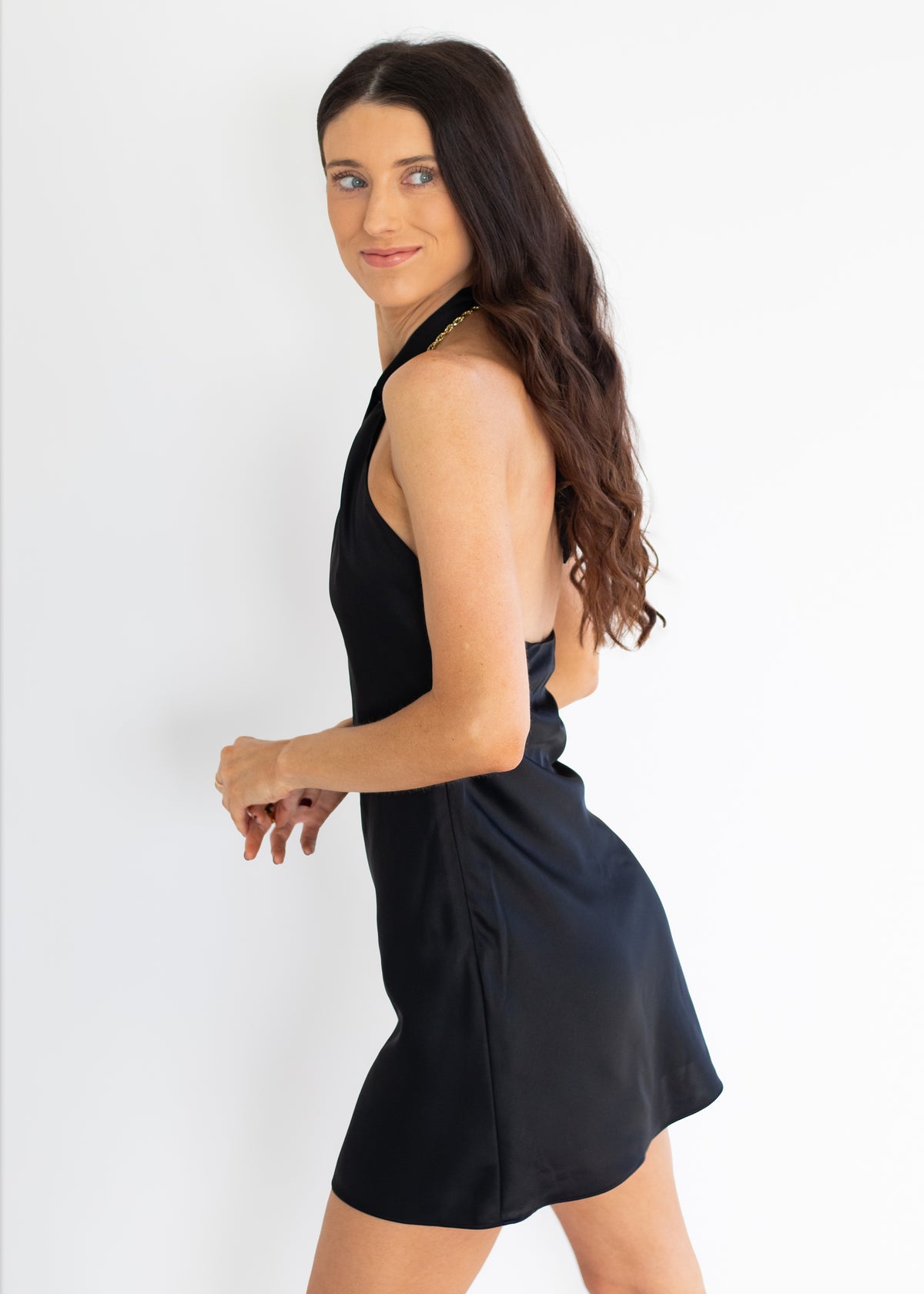 A woman modeling a sleeveless black halter mini dress with a flared skirt, photographed against a plain white studio background.