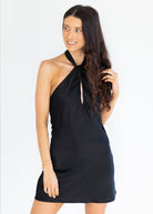 A woman modeling a black halter-neck mini dress with a tie-front keyhole detail, photographed against a clean white studio background.