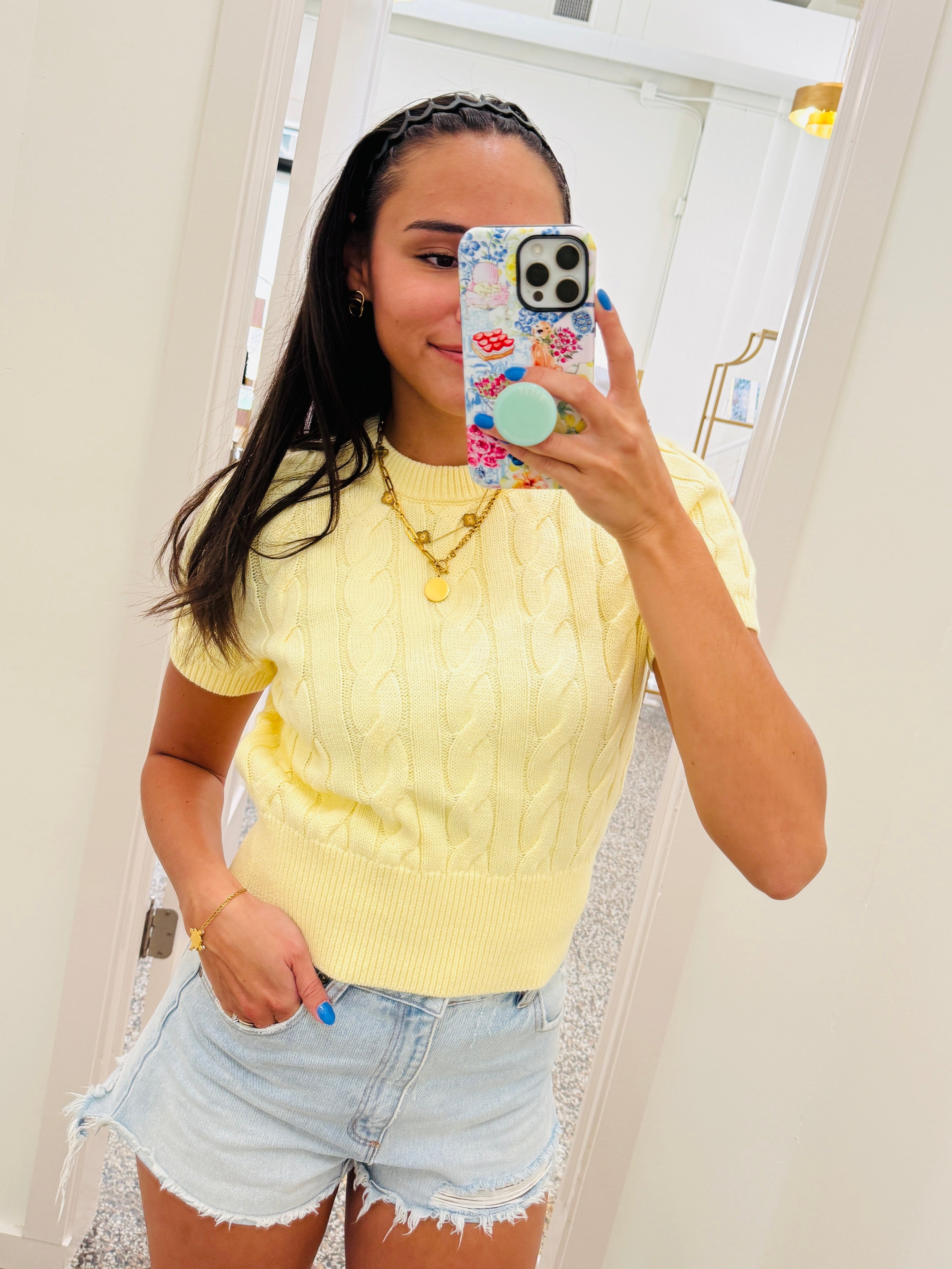 A woman takes a mirror selfie wearing a pale yellow short-sleeve cable-knit top and light-wash denim shorts in a brightly lit indoor room.