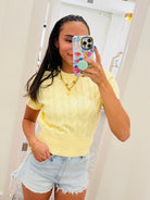 A woman takes a mirror selfie wearing a pale yellow short-sleeve cable-knit top and light-wash denim shorts in a brightly lit indoor room.