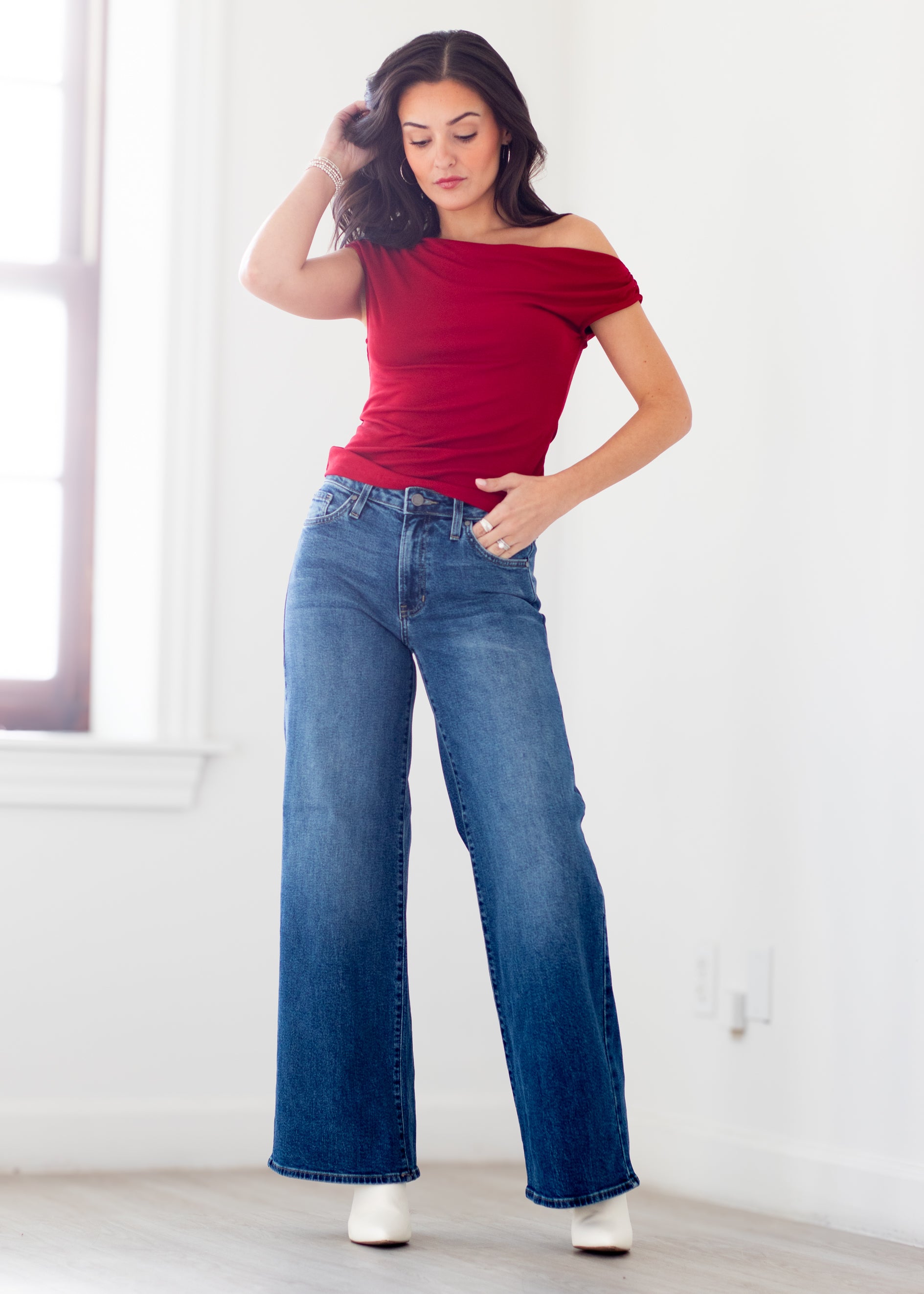 A woman modeling a red off-the-shoulder short-sleeve top and blue wide-leg jeans in a bright, white-walled room.