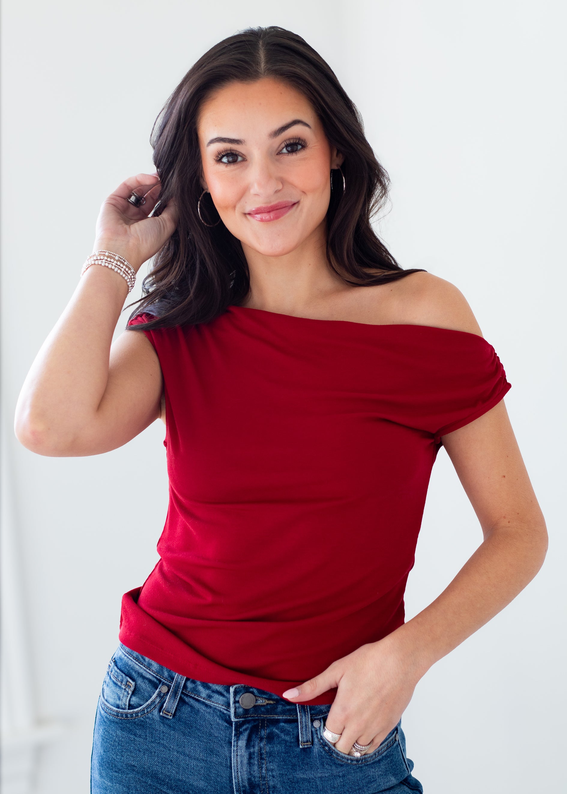 A woman modeling a red off-the-shoulder short-sleeve top with blue jeans, posing against a clean white background.