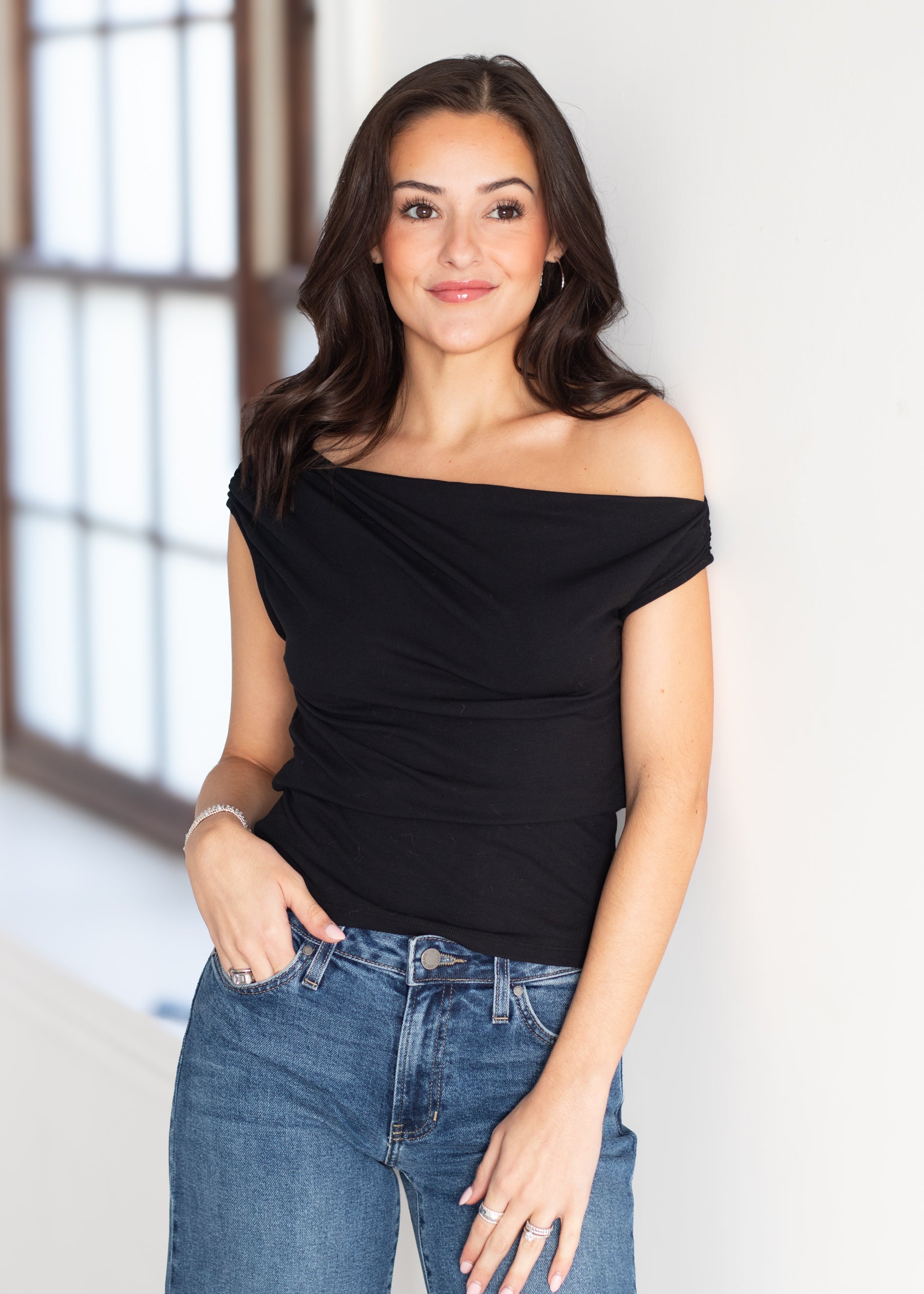 A woman modeling a black off-the-shoulder draped top with blue jeans, standing in a brightly lit room with a white wall and a window in the background.