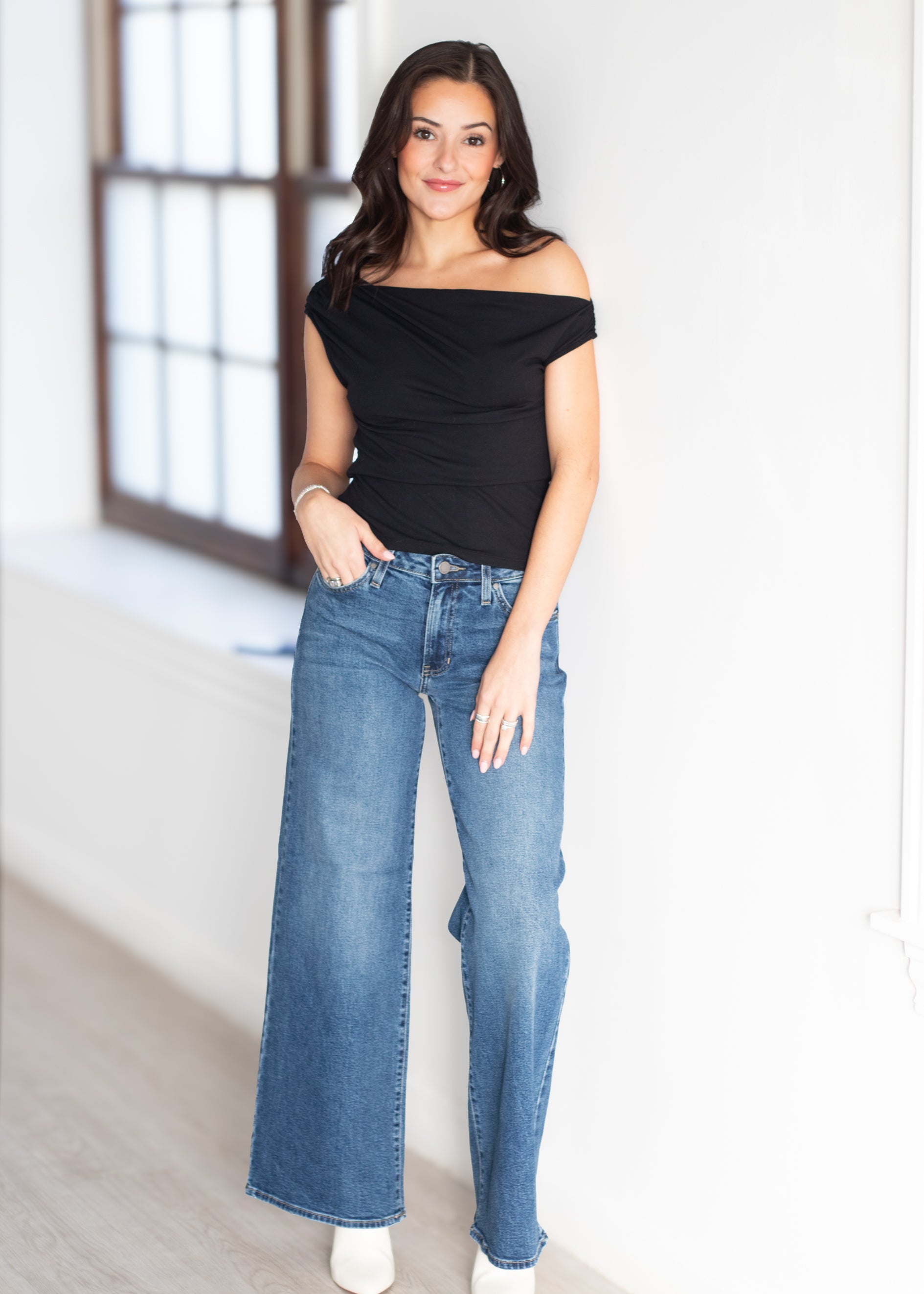 A woman modeling a black off-the-shoulder ruched top and high-waisted, wide-leg blue jeans in a bright, modern room with a large window.