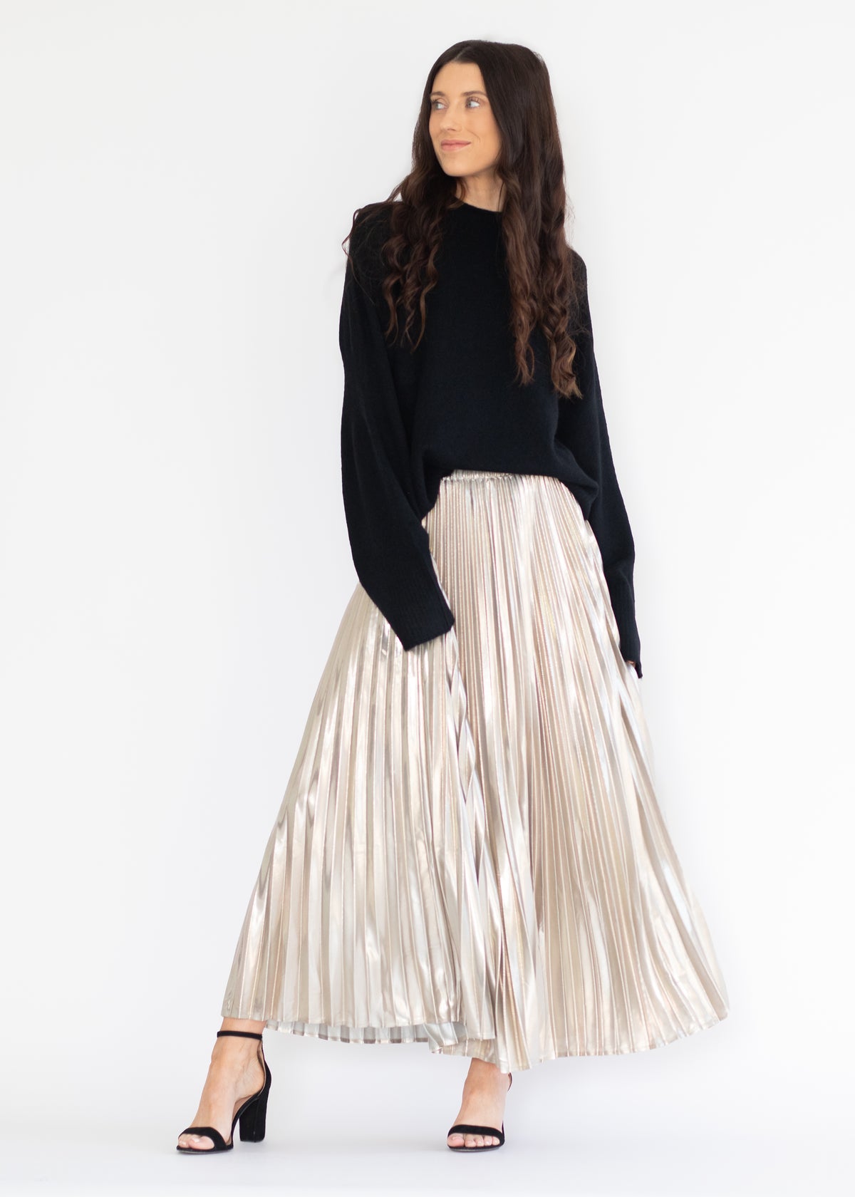 A woman modeling a black knit long-sleeve top and a high-waisted, metallic champagne pleated maxi skirt, paired with black heeled sandals, against a plain white studio background.