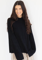 A woman with long, wavy brown hair wearing an oversized black knit mock-neck sweater and a pleated light-colored skirt, posing against a plain white studio background.