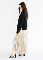 A woman in a black cardigan and a shiny pleated champagne-colored maxi skirt, standing in profile against a plain white background.
